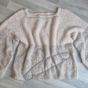 May include: A light beige knitted sweater with long, voluminous sleeves and a square neckline. The sweater is lying flat, with a silver metal hanger resting on top. The knitwear has a textured appearance.