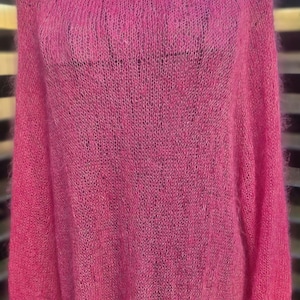 May include: A bright pink, open-weave sweater with a wide neckline and long sleeves. The knitwear is displayed on a black metal hanger against a blurred background. The sweater's texture appears soft and slightly sheer.
