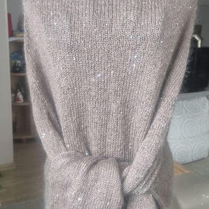 May include: A taupe-colored knitted wrap with a sparkly, sequined design. The wrap is tied in the front, creating a draped effect. The knitwear is displayed on a mannequin, showcasing its texture and style. This elegant piece is suitable for special occasions.