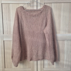 May include: A dusty rose-colored sweater with a round neckline and long sleeves, hanging on a silver hanger. The knitwear has a soft, textured appearance. The sweater is displayed against a light wood-grain background.