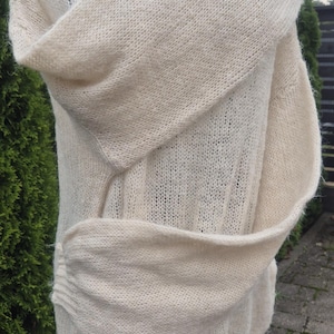 May include: Cream-colored, open-weave knit shawl or wrap. The garment is draped over a form, showcasing its soft texture and flowing design. The shawl appears to be made of a lightweight, possibly wool or a wool blend, material.