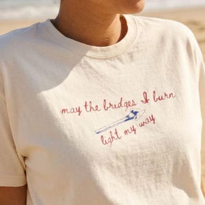 May include: Cream-colored t-shirt with the embroidered phrase "may the bridges I burn light my way" in red and blue. The shirt is paired with navy blue bottoms, creating a casual beach-ready look.