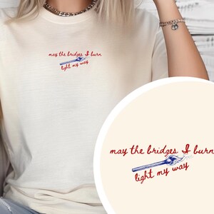 May include: A cream-coloured t-shirt featuring the phrase "may the bridges I burn light my way" in red and blue. The design includes a burning match. A casual, everyday wear item.