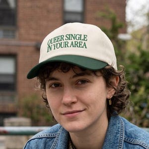 Op de afbeelding: Een baseballpet met een gebroken witte kroon en een donkergroene klep. Op de voorkant van de pet staat de tekst "QUEER SINGLE IN YOUR AREA" geborduurd in donkergroene hoofdletters. De pet heeft een gestructureerd voorpaneel en een gebogen klep.