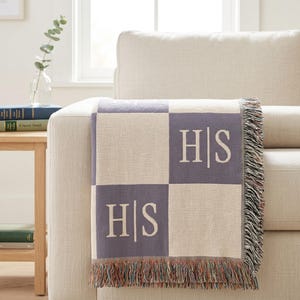 Puede incluir: Una manta tejida con un patrón de tablero de ajedrez en gris claro y lavanda. La manta presenta las iniciales "HS" en blanco en dos de los cuadrados. La manta tiene un borde con flecos y está colocada sobre un sofá beige.