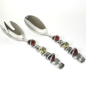 Puede incluir: Un juego de dos utensilios para servir ensalada de plata con cuentas de vidrio rojas y amarillas envueltas alrededor de los mangos.