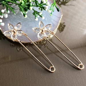 Può includere: Due spille di sicurezza dorate con accenti di fiori di cristallo trasparente. Le spille sono su uno sfondo grigio chiaro.