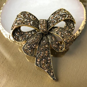 Pode incluir: Broche de laço em estilo vintage, em ouro antigo. O laço é detalhado com um design floral em filigrana e adornado com pequenos cristais âmbar brilhantes. O broche está sobre um fundo neutro.