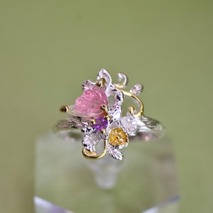 May include: A silver ring with a floral design, featuring a pink gemstone, purple and yellow accents. The ring has gold-colored details and is set against a soft green background.