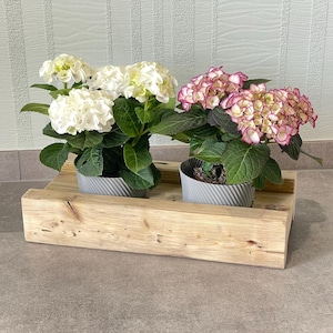 Könnte beinhalten: Zwei Topf-Hortensien, eine mit weißen Blüten und die andere mit rosa und weißen Blüten, stehen auf einem rustikalen Holzpflanzer. Der Pflanzer ist aus hellem Holz gefertigt und hat ein einfaches, rechteckiges Design.