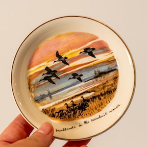 May include: A small, round, off-white decorative plate with a gold rim. The plate features a painted landscape with flying ducks against a sunset sky. Text at the bottom reads "Mallards in the windmill marsh."