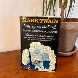 May include: A hardcover book titled "Letters from the Earth" by Mark Twain, edited by Bernard DeVoto. The book has a black cover with yellow and white text and a blue and white illustration of a man's face. The book is on a wooden surface.