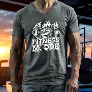 May include: Gray V-neck t-shirt with a white graphic. The graphic features a weightlifting barbell held by a fist, flames, and the words "FITNESS MODE ON". The shirt is made of a soft material.