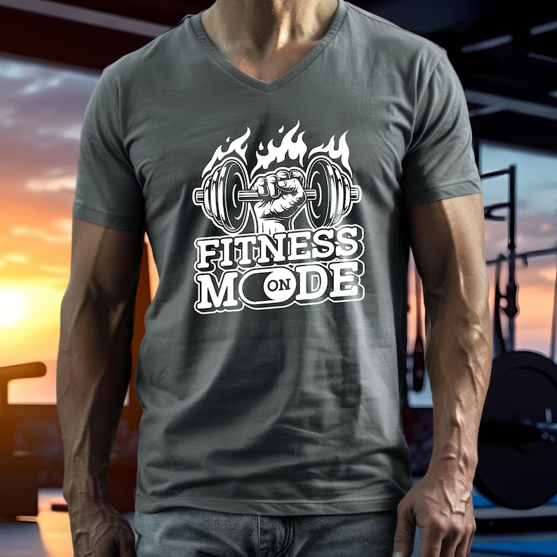May include: Gray V-neck t-shirt with a white graphic. The graphic features a weightlifting barbell held by a fist, flames, and the words "FITNESS MODE ON". The shirt is made of a soft material.