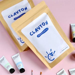 Könnte beinhalten: Zwei beige Beutel mit dem Wort "CLAYTON" in Blau und dem Text "Selbsttrocknender Ton" auf einem weißen Etikett. Darunter eine blaue sonnenähnliche Grafik. Tuben Acrylfarbe in verschiedenen Farben sind sichtbar.