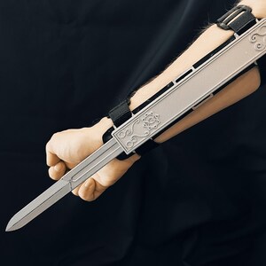 Könnte beinhalten: Eine silberfarbene versteckte Klingen-Requisite mit dekorativen floralen und scrollartigen Details. Die Klinge ist an einer schwarzen Handgelenk- und Unterarmstütze mit verstellbaren Riemen befestigt. Die Requisite ist für das Tragen am Unterarm konzipiert.