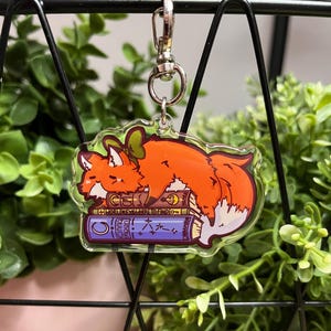 Könnte beinhalten: Ein klarer Acryl-Schlüsselanhänger mit einem Cartoon-Fuchs mit orangefarbenem Fell und einer grünen Schleife, der auf einem Stapel Bücher schläft. Die Bücher tragen den Text "CE" und "WORLD KEYS & WHERE TO FIND ON". Der Schlüsselanhänger hat einen silbernen Verschluss.