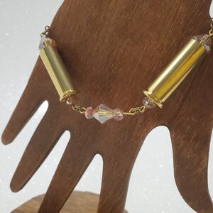 May include: A gold-colored bracelet featuring two brass bullet casings connected by a delicate chain. The chain incorporates small, faceted, pink and clear beads, adding a touch of sparkle to the design. The bracelet is displayed on a wooden hand-shaped stand.