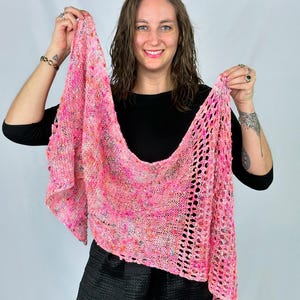 Puede incluir: Un chal de punto rosa y multicolor con un diseño calado similar al encaje. El chal se sostiene, mostrando sus colores vibrantes y su patrón texturizado. La paleta de colores incluye tonos de rosa, naranja y amarillo.