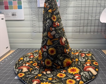 May include: A quilted witch hat featuring a black fabric base with a repeating pattern of yellow sunflowers, various mushrooms, and green foliage. The hat has a tall, pointed crown and a wide, circular brim, with visible stitching throughout the fabric.