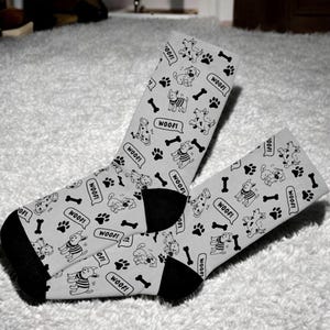 May include: A pair of gray socks with a black and white pattern of dogs and bones. The socks have the word "WOOF!" printed on them.