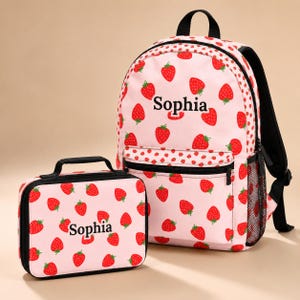 Puede incluir: Una mochila rosa claro y una bolsa de almuerzo rectangular a juego, ambas con un patrón repetitivo de fresas rojas. Cada artículo está personalizado con el nombre Sophia impreso en negro. La mochila incluye un bolsillo frontal con cremallera.