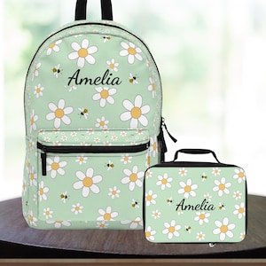 May include: A light green backpack and matching lunch bag set. Both items feature a daisy and bee pattern with the name "Amelia" printed in black. The backpack has a front pocket and black zipper.