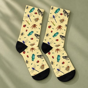 May include: A pair of crew socks with a light yellow background featuring a repeating pattern of various insects including blue dragonflies, red ladybugs, brown spiders, striped beetles, green grasshoppers, and black and white moths with black cuffs.