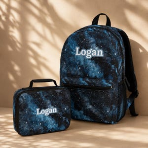 May include: A backpack and a matching lunch box, both featuring a dark blue and black galaxy print with white stars. Each item is personalized with the name Logan printed in a light blue font on the front. The backpack has a top handle and side pockets.