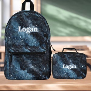 May include: A backpack and matching lunch bag set. Both items feature a dark blue and black galaxy print with white speckles. The name "Logan" is printed in white on both the backpack and lunch bag. The backpack has a black zipper and straps.