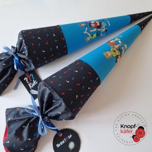 Könnte beinhalten: Zwei blaue und schwarze Stoff-Schultüten mit Comicfiguren. Die Tüten haben einen schwarzen Stoffabschnitt mit weißen, roten und blauen Fragezeichen. Jede Tüte ist mit einem blauen Band gebunden und hat ein Etikett mit der Aufschrift "??? Kids".