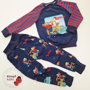 Könnte beinhalten: Marineblaues zweiteiliges Kinder-Schlafanzug-Set. Das langärmlige Oberteil hat rot-blau gestreifte Ärmel und eine Grafik eines Elefanten, der einen Bulldozer fährt. Die passende Hose zeigt das gleiche Elefanten- und Bulldozer-Design.