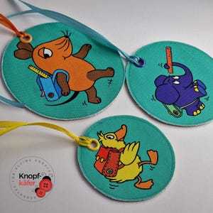 Könnte beinhalten: Drei runde, türkisfarbene Stoffanhänger mit bunten Tierillustrationen. Jeder Anhänger hat oben einen kleinen, runden Metallring. Die Tiere sind eine Maus, ein Elefant und eine Ente. Jedes Tier trägt einen kleinen, roten Rucksack. Der Text "Knopf-käfer" steht auf einem der Anhänger.