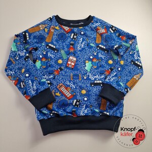 Könnte beinhalten: Blaues Sweatshirt mit London-Motiv. Das Design umfasst Illustrationen von Bussen, Big Ben, Telefonzellen und dem Buckingham Palace. Das Sweatshirt hat marineblaue Bündchen und einen marineblauen Ausschnitt.
