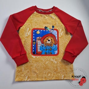 Könnte beinhalten: Ein langärmeliges Shirt mit roten Ärmeln und einem senfgelben Körper. Der Körper hat einen London-Themen-Print und einen Aufnäher mit einem Cartoon-Bären, der ein Sandwich isst. Der Bär trägt einen blauen Mantel und einen roten Hut. Ein Vogel sitzt über dem Bären.