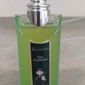Puede incluir: Un frasco de perfume de vidrio verde con tapón plateado. El frasco tiene una etiqueta rectangular con la marca "BVLGARI" y las palabras "Eau Parfumée" y "au thé vert EXTREME". El perfume en su interior es de color verde claro.