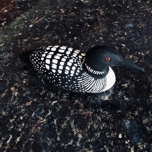 May include: A wooden decorative loon figurine. The bird has a black head and beak, with a white throat and chest. The back is black with white polka dots. The loon has red eyes and is resting on a dark surface.