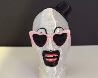 May include: A white 3D printed figurine of a person wearing a black top hat, pink heart-shaped sunglasses, and a black mask with a red mouth and white teeth.