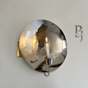 May include: A silver wall sconce with a round, hammered metal shade and a single candle holder. The candle is lit and the flame is visible.