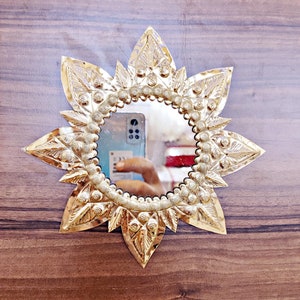 Puede incluir: Un espejo de metal dorado con un diseño decorativo en forma de estrella. El espejo es redondo y está rodeado por un borde de pequeños círculos en relieve.