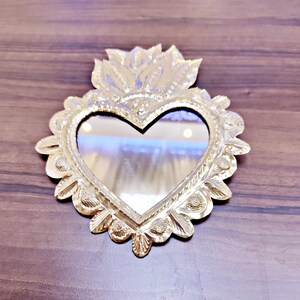 以下が含まれることがあります： 縁に装飾的な花のデザインが施された、金色のハート型の鏡。鏡は青と白の背景を反射している。