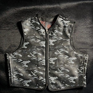 Könnte beinhalten: Schwarze Camouflage-Weste mit durchgehendem Reißverschluss. Die Weste hat ein perforiertes Design und ein Camouflagemuster in Grau- und Schwarztönen. Die Weste hat einen V-Ausschnitt und eine schwarze Einfassung.