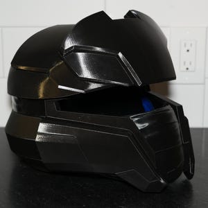 May include: A black plastic helmet with a raised visor. The helmet has a textured surface and a sleek, futuristic design.