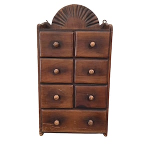 May include: A small, vintage wooden cabinet with eight drawers and a decorative sunburst design at the top. The cabinet is made of dark brown wood and has small, round knobs on each drawer. It is designed to be mounted on a wall.