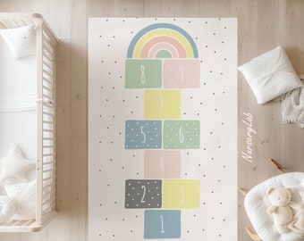 May include: A white play mat with a rainbow and a hopscotch game with numbers 1 through 9. The hopscotch squares are in different colors, including green, blue, yellow, pink, and gray. The mat has a white background with small black dots.