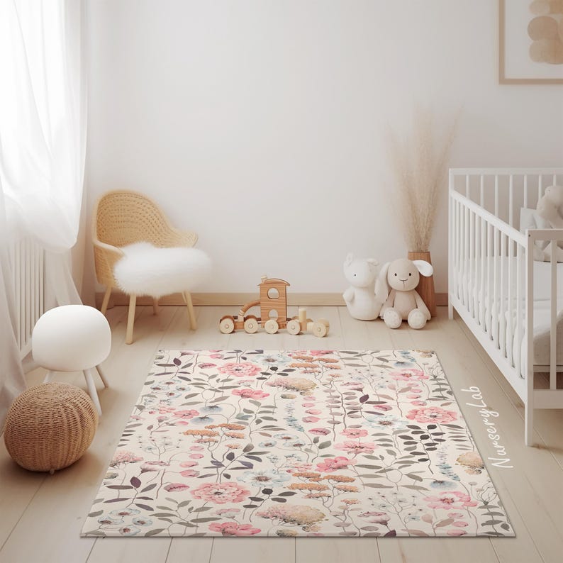 May include: A rectangular rug with a floral pattern in pink, beige, and gray tones. The rug features a variety of flowers and leaves, creating a soft and inviting look. The rug is placed in a nursery setting, with a crib and toys visible.