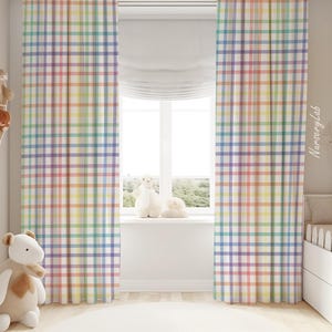 Op de afbeelding: Een paar regenboog ruitgordijnen met een witte achtergrond. De gordijnen hangen in een kinderkamer met een wit ledikant en knuffels.