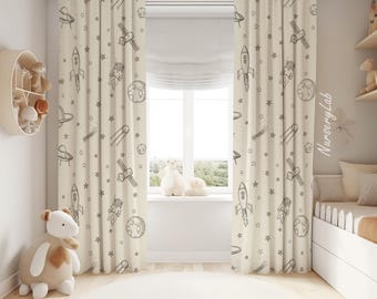 May include: Off-white curtains featuring a repeating pattern of dark gray space-themed illustrations including rockets, astronauts, planets, satellites, and stars. The fabric hangs in vertical folds, covering a window in a room with light walls.