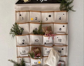 Op de afbeelding: Een witte stoffen adventskalender met 24 vakjes, genummerd van 1 tot en met 24. De kalender is in de vorm van een huis met een zwart dak en een zwarte ster op het dak. De naam "Myla" staat op het dak. In sommige vakjes zitten kleine cadeautjes.
