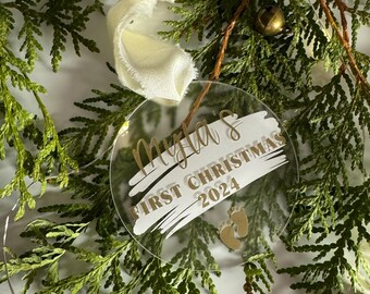 May include: A clear round ornament with gold lettering that reads "Myla's First Christmas 2024" and a gold footprint design. The ornament is hanging from a white ribbon and is surrounded by green pine branches.